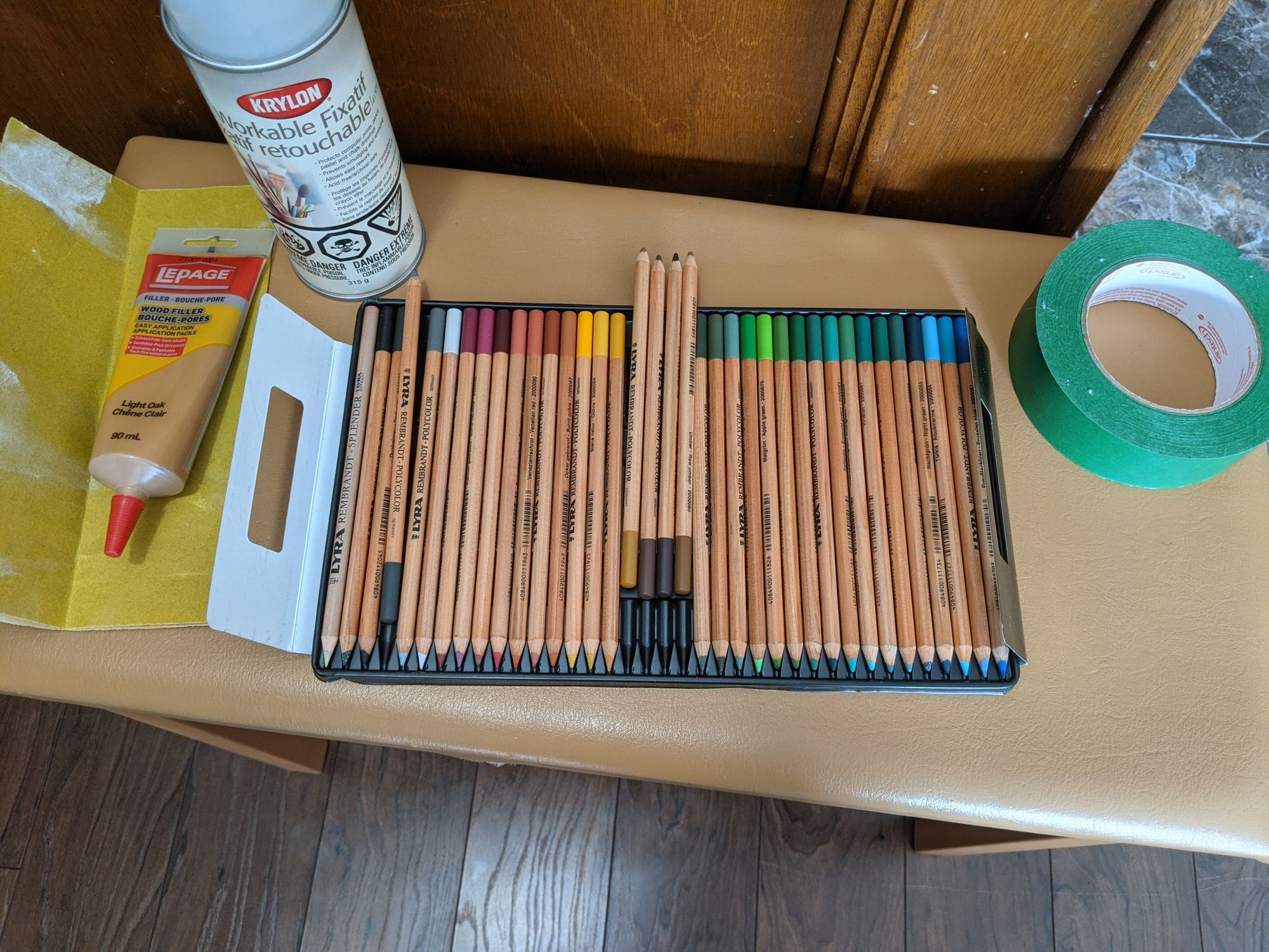 Sandpaper, wood filler, workable fixative, pencil crayons, and masking tape displayed on a piano bench.
